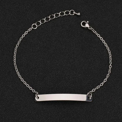 Silver thin chain bracelet with a plain rectangular nameplate and extension chain, on a black textured background.