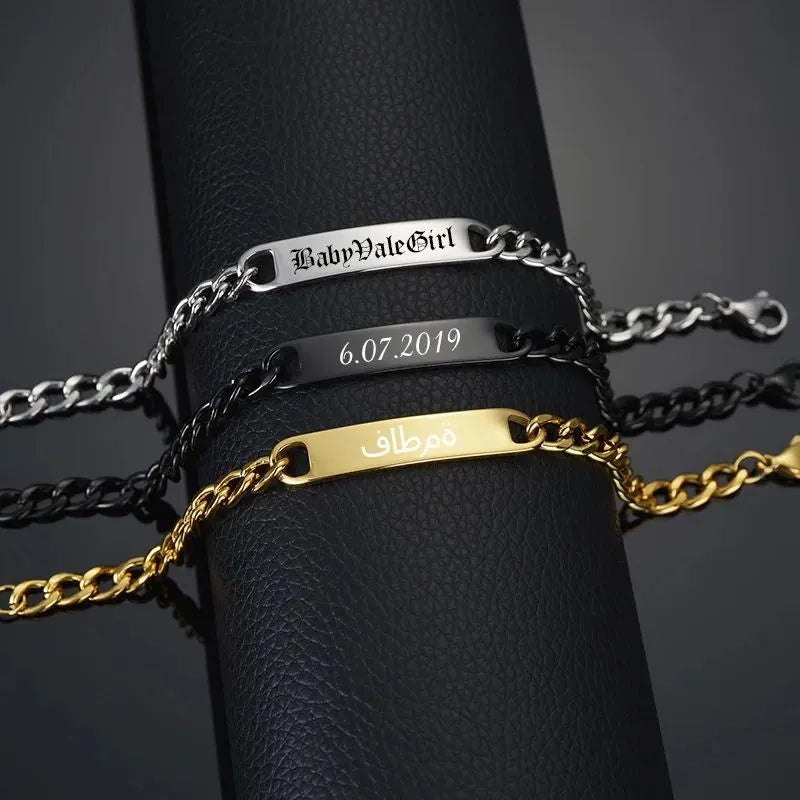 Silver, black and gold nameplate bracelets draped over a black leather stand, each engraved with a different sample name, date and script