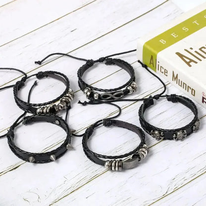 Five individual black leather bracelets from the Rivet and Infinity set, including a studded band and an infinity charm, laid out on a wooden surface with a book.