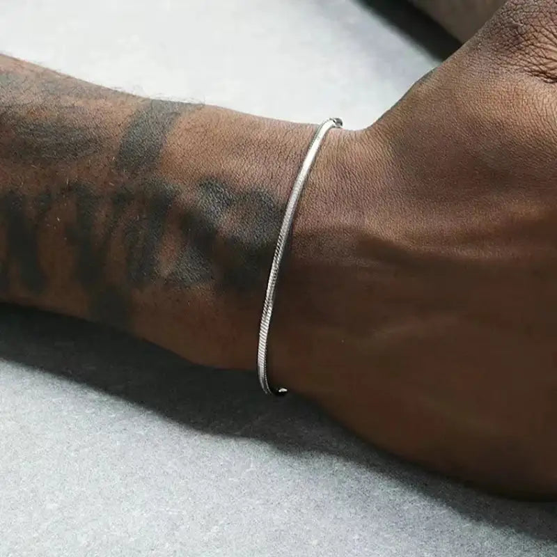 Silver round snake chain bracelet worn on the wrist, resting on a light grey surface