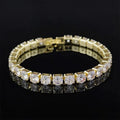 Gold tennis bracelet with square-set round cubic zirconia stones laid in a loop on a black background, clasp visible and stones reflected below.