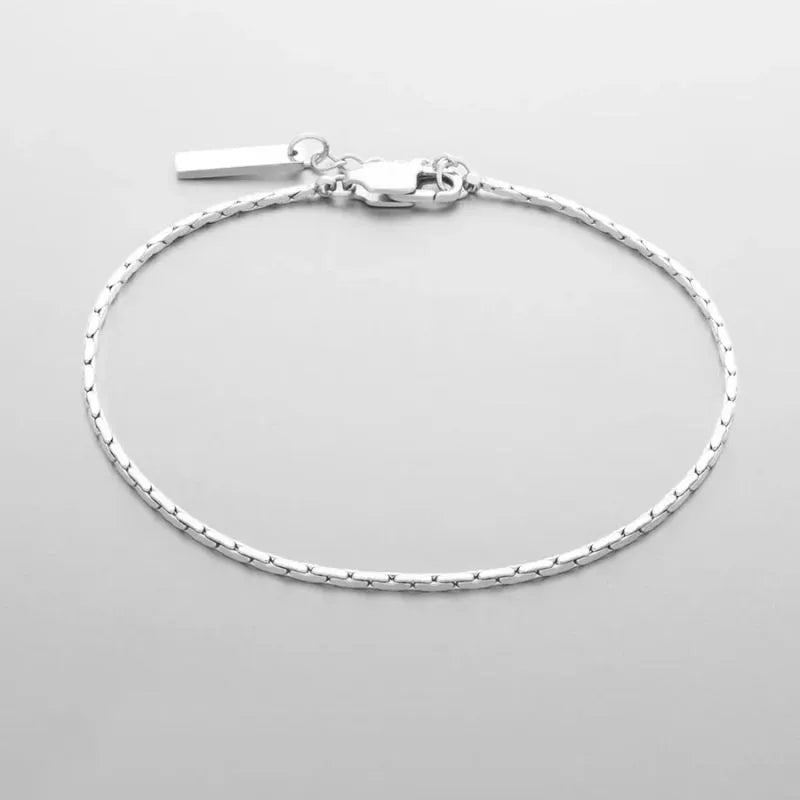Silver-tone wire chain bracelet with a rectangular bar tag charm and lobster clasp, laid in an oval on a light grey background.