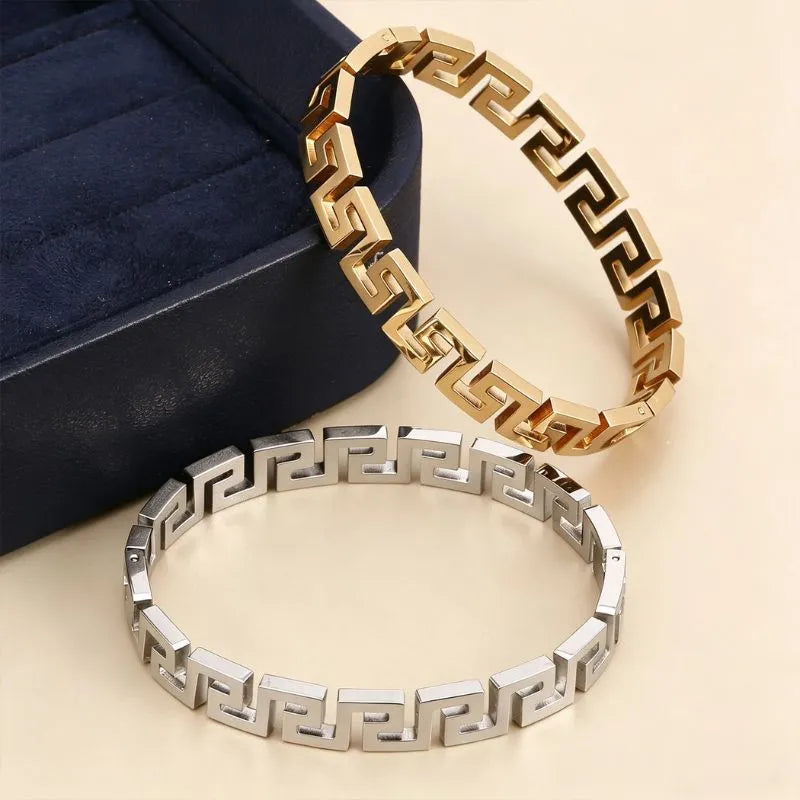 Gold and silver Greek key bangle bracelets resting together beside a navy blue jewellery box on a beige surface