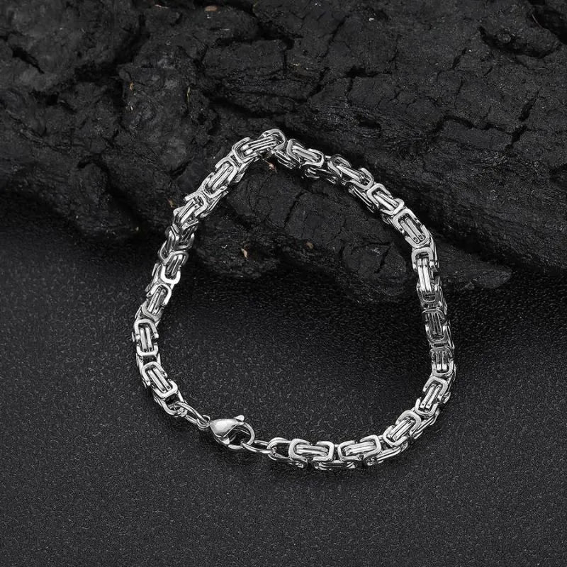 Silver-tone Byzantine box-chain bracelet laid flat in a loop against a dark, charcoal-textured backdrop.