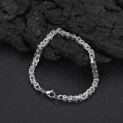 Silver-tone Byzantine box-chain bracelet laid flat in a loop against a dark, charcoal-textured backdrop.