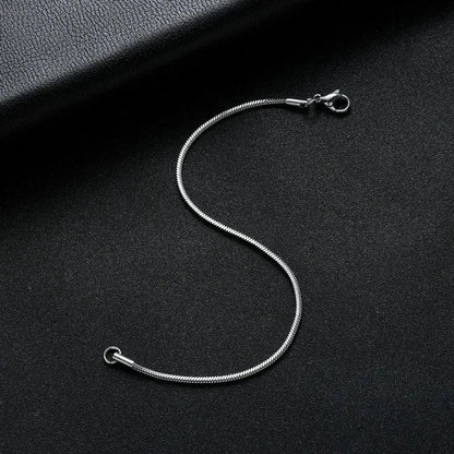 Silver snake chain bracelet laid in an S-curve on a black leather surface, with a dark leather book prop in the corner.