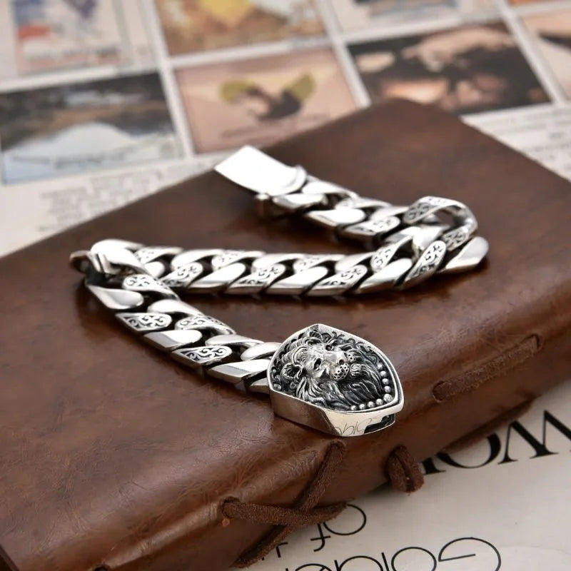 Silver Cuban-link bracelet with a lion-head shield centerpiece shown open, draped over a leather journal with a postcard-style photo collage in the background.