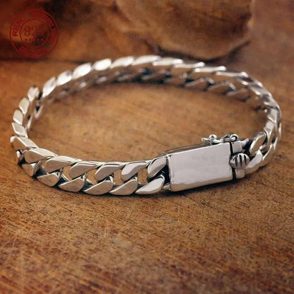 Silver Cuban-link bracelet laid in an oval on a textured wooden surface, with a stone prop and red sterling silver hallmark stamp visible.